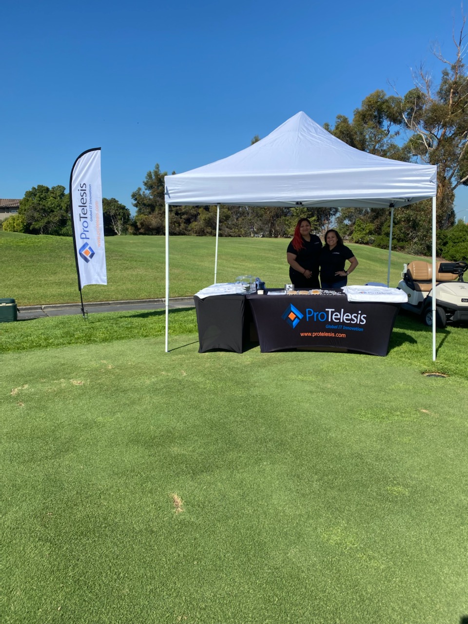 ProTelesis team standing ath the company stand at the San Ysidro Health's 16th Annual Clasico de Golf tournament, with the logo falg in frame.