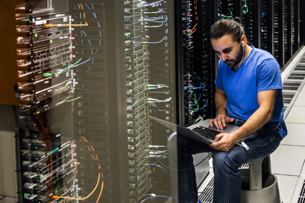 A man sitting on a stool working on his laptop in a server room